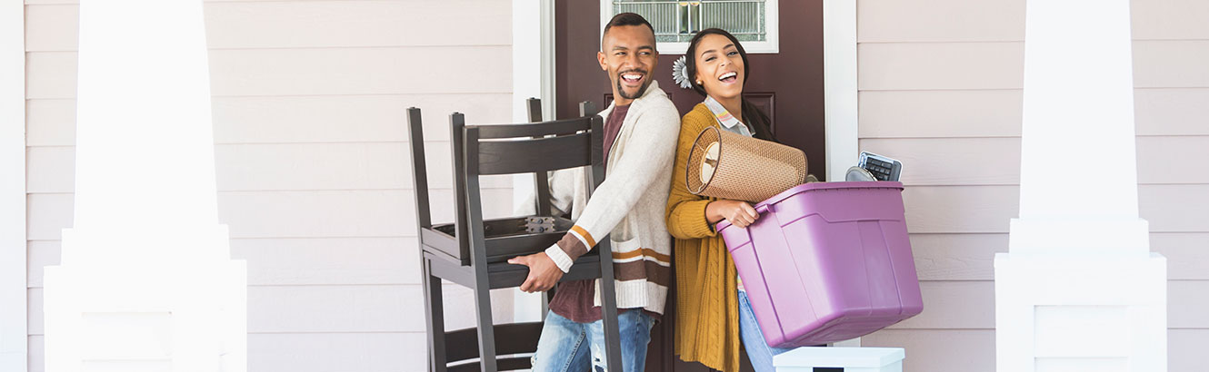 Man and woman on front porch carrying items into new home.